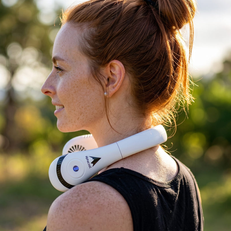 Woman with freckles and red hair in a bun wears white neck fan with "elbha cool" logo, outdoors in warm sunlight.