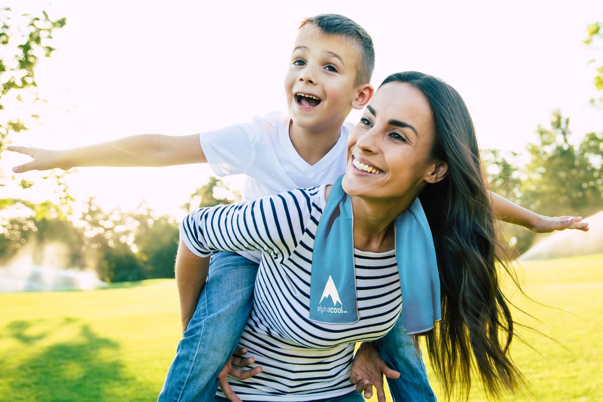 Mother giving son piggyback ride in sunny park, both smiling, blue towel with "alphacool" logo draped around her neck.